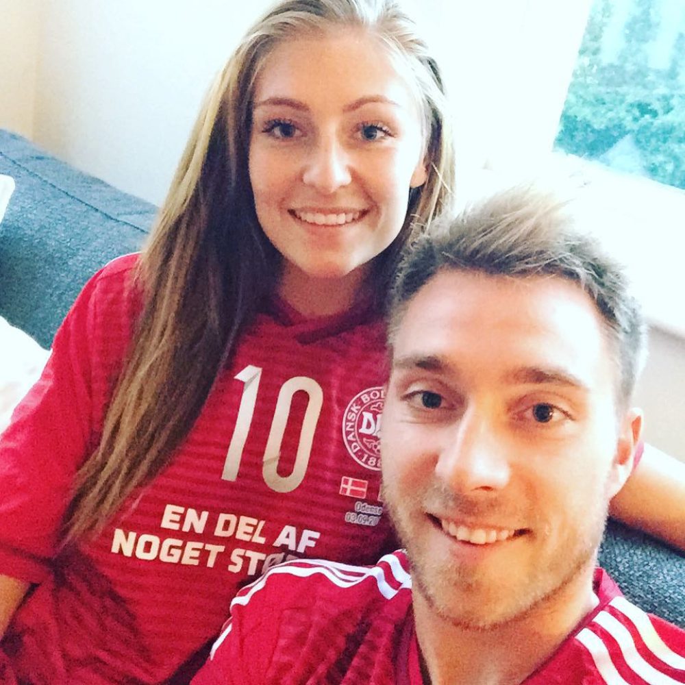 Fußball-Profi Christian Eriksen und seine Ehefrau Sabrina Kvist Jensen tragen das Dänemarktrikot und lächeln in die Kamera.